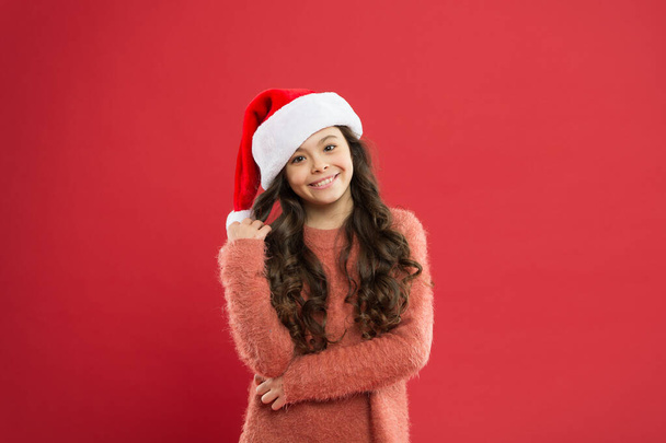 Playful cutie. Adorable girl with long curly hair wear santa claus hat red background. Counting days till christmas. Christmas party for kids and students. Christmas holiday invitation concept - Photo, Image