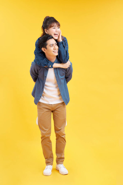 Image of beautiful couple man and woman in basic clothes smiling together while doing piggyback ride isolated over yellow background - Photo, Image