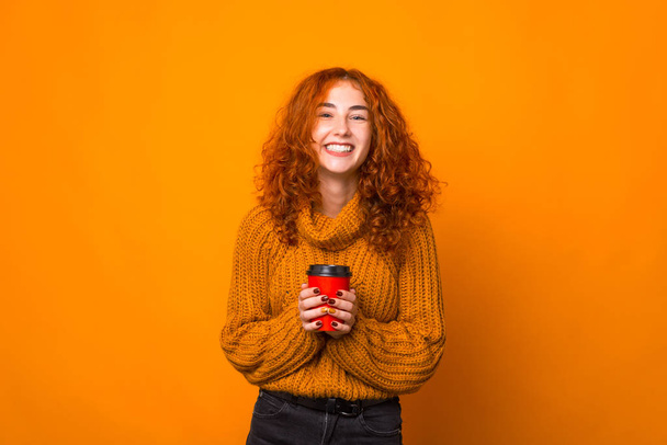 Photo of reddish girl, smiling at camera and holding a cup of coffee or tea, over orange background - Foto, afbeelding