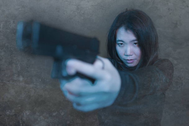 Asian girl pointing handgun - dramatic action portrait of young attractive and dangerous looking Chinese woman holding gun firing in dark grunge background - Photo, Image