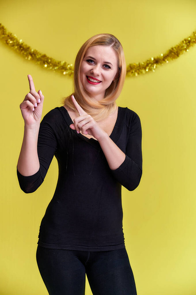 Concept of a young blonde woman with New Year's decor. Portrait of a cute girl in a black T-shirt with long beautiful hair and great makeup. Smiling, showing emotions on a yellow background. - Fotó, kép