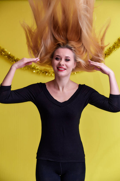 Concept of a young blonde woman with New Year's decor. Portrait of a cute girl in a black T-shirt with long beautiful hair and great makeup. Smiling, showing emotions on a yellow background. - Foto, Imagen