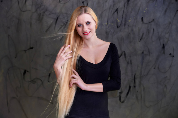 Concept of a young blonde woman with a chic hairstyle. Portrait of a cute girl in a black T-shirt with long beautiful hair and great makeup. Smiling, showing emotions on a gray background. - Foto, Imagem