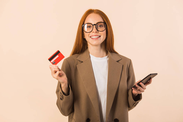 beautiful young woman with red hair in a jacket on a beige background with a credit card and a phone in her hands - Foto, Imagem