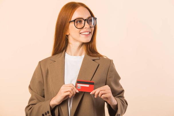 beautiful young woman with red hair in a jacket on a beige background with a credit card in hands - Photo, image