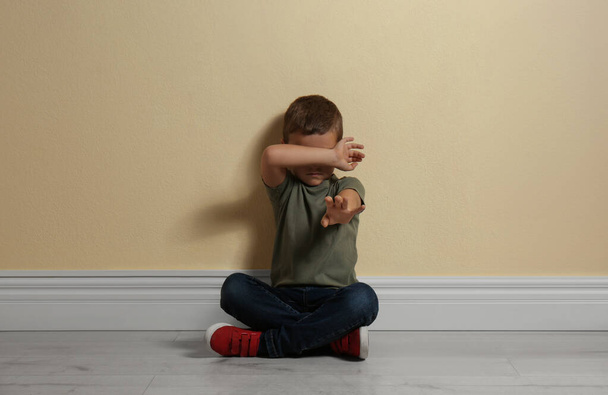 Scared little boy closing face with hand on floor near yellow wall. Child in danger - Foto, afbeelding