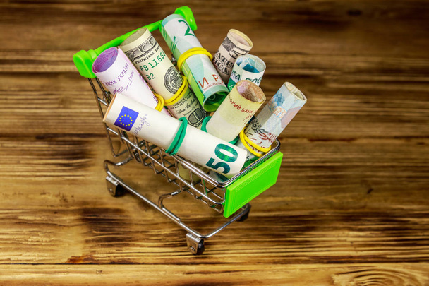 Shopping cart with money from different countries: euros, us dollars, ukrainian hryvnia, russian rubles, egyptian pounds on wooden background - Foto, afbeelding