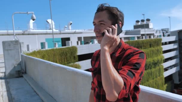 Улыбающийся молодой человек в клетчатой рубашке разговаривает по мобильному телефону на крыше здания на фоне города
 - Кадры, видео