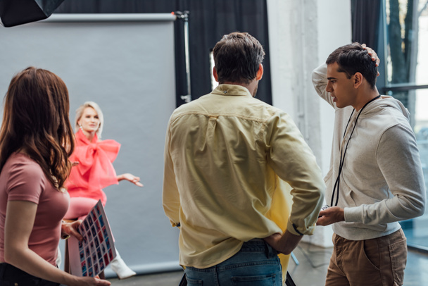 πίσω όψη του καλλιτέχνη σκηνοθέτη στέκεται με το χέρι στο ισχίο κοντά στους συναδέλφους και το μοντέλο στο στούντιο φωτογραφιών  - Φωτογραφία, εικόνα