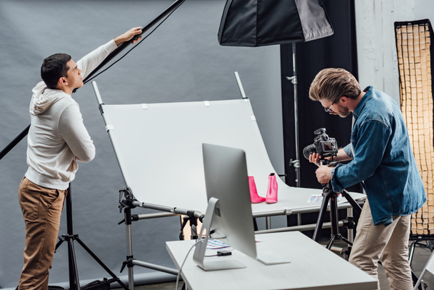 handsome assistant touching reflector while photographer photo shooting shoes  - Foto, imagen