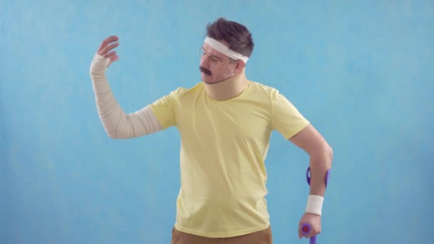 Funny mustachioed man after a sports injury with crutches poses for the camera standing on a blue background - Séquence, vidéo