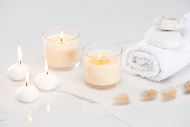 flauschiges Hasenschwanzgras in der Nähe brennender weißer Kerzen im Glas und gerolltem Handtuch mit Steinen auf marmorweißer Oberfläche - Foto, Bild
