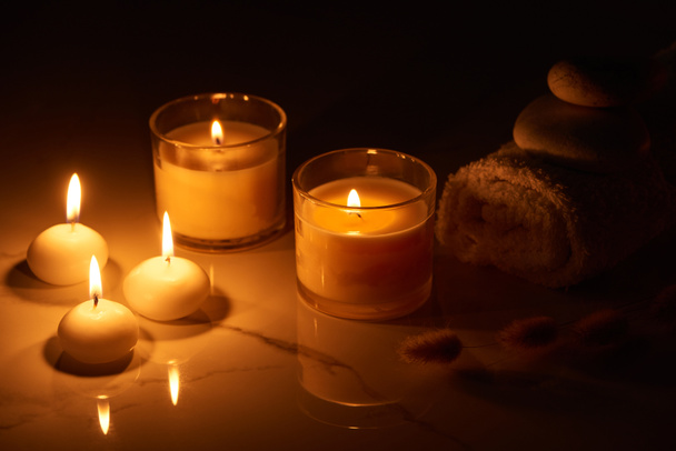 burning candles in glass glowing in dark near rolled towel on marble surface - Photo, Image