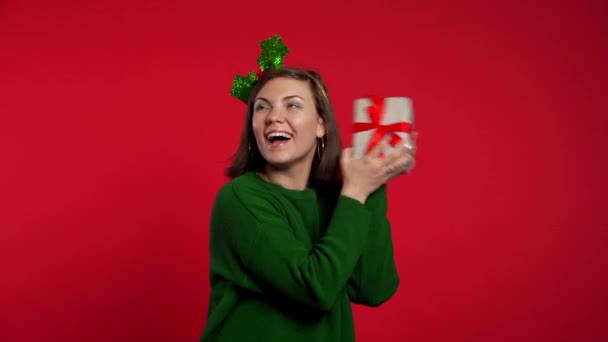 Young woman smiling and holding gift box near ear to guess whats inside on red studio background. Girl in Santa hat. Christmas mood. - Video