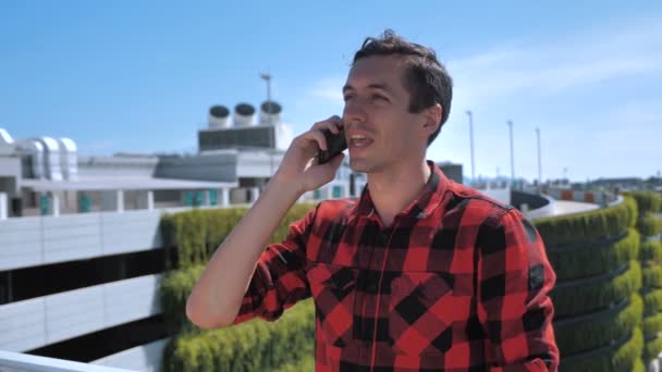 Glimlachende jonge man praat mobiele telefoon buiten op het dak. Portret van ambitieuze zakenman die buiten over mobiele telefoon praat op stedelijke achtergrond. Ondernemer bespreken van zaken via de telefoon buiten. - Video