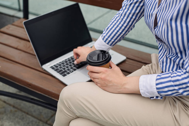 A girl sits on a bench in city with a cup of takeaway coffee and opened laptop - Foto, Imagem