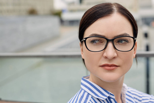 Senhora de negócios concentrada grave com cabelos escuros e olhos azuis usando óculos em uma camisa listrada olha para o lado
 - Foto, Imagem