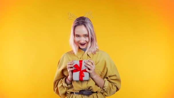 Unusual woman with pink hair smiling and gives gift, hands it to the camera on yellow studio background. Hipster girls portrait in trendy outfit. Christmas mood. - Filmagem, Vídeo