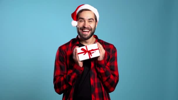 Young handsome man smiling and holding gift box on blue studio background. Guy in Santa hat. New year mood - Filmagem, Vídeo