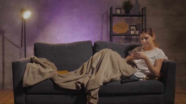Młoda piękna dziewczyna leżąca na kanapie i pisząca wiadomości przez telefon - Materiał filmowy, wideo
