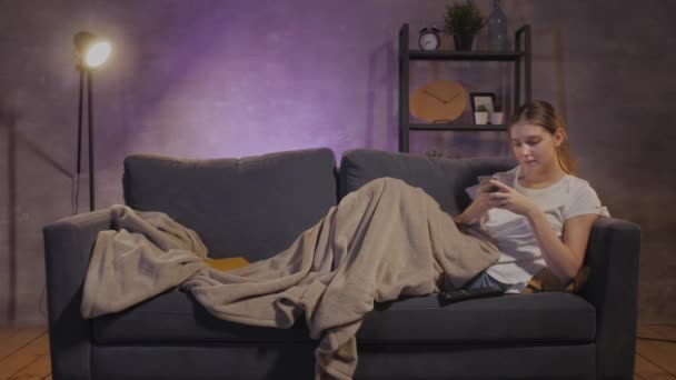 Nuori kaunis tyttö makaa sohvalla ja kirjoittaa viestejä puhelimeen
 - Materiaali, video
