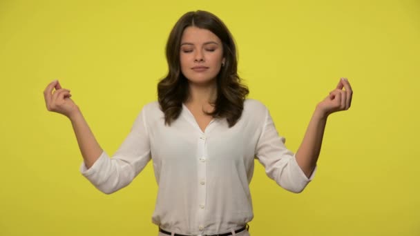 Peaceful girl with brunette hair in blouse holding fingers in mudra gesture, meditating practicing mindful yoga, having calm concentrated expression. indoor studio shot isolated on yellow background - Metraje, vídeo