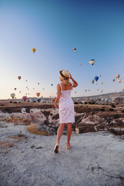 Jeune femme en robe et chapeau sur un sommet de montagne bénéficiant d'une vue magnifique sur le lever du soleil et les ballons en Cappadoce. - Photo, image