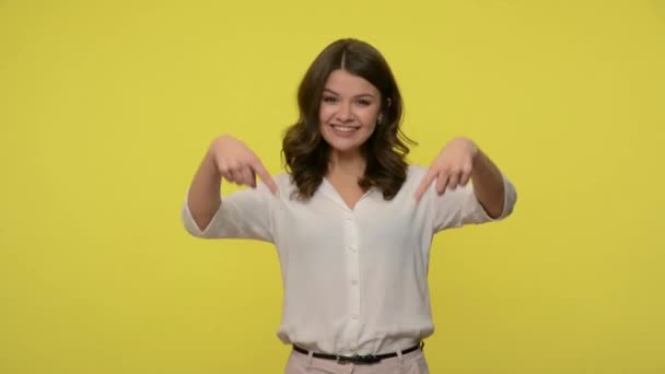 Friendly cheerful brunette woman pointing down and smiling at camera, meaning subscribe or follow information below, showing place for promotion text, advertising area. studio shot, yellow background - Filmagem, Vídeo