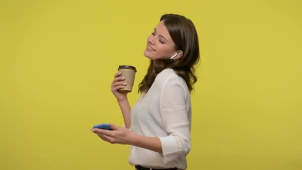 Cheerful energetic woman with brunette hair in blouse holding mobile phone and cup of coffee, listening to music in earplugs and dancing, smiling at camera. studio shot isolated on yellow background - Séquence, vidéo