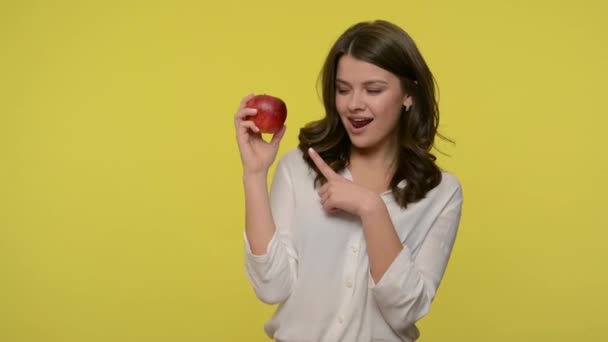 Beautiful slim woman pointing at red apple in her hand and looking seductively to camera, showing thumbs up, recommending fruit, healthy delicious diet food. studio shot isolated on yellow background - Кадри, відео