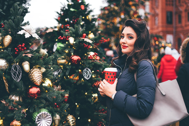 Smiling woman with paper cup near Christmas trees looks at view. - Zdjęcie, obraz