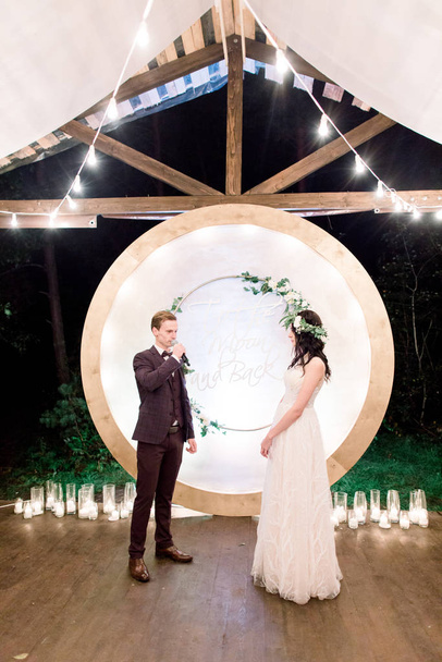 夜の結婚式を屋外で花嫁と新郎は結婚式のアーチの背景にお互いの誓いを誓う。美しい夫婦だ電球付きの素朴な木造レストラン - 写真・画像