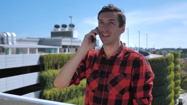 Glimlachende jongeman in een geruit hemd aan het praten op een mobiele telefoon op het dak van een gebouw tegen de achtergrond van de stad - Video