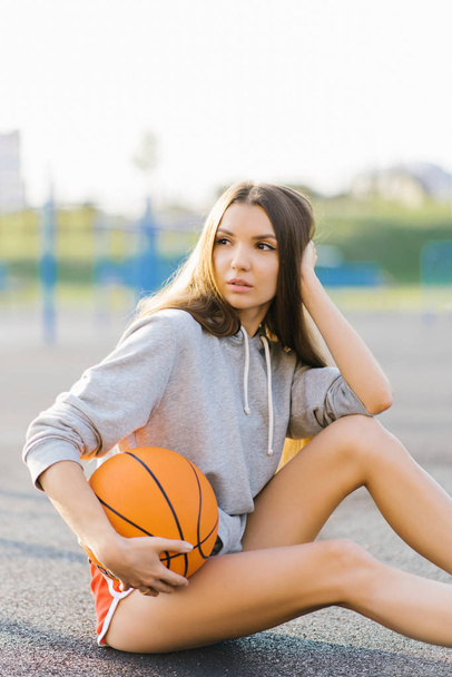 Κορίτσι μπασκετμπολίστας κάθεται στην παιδική χαρά και κρατώντας την μπάλα στα χέρια του και κοιτάζει μακριά - Φωτογραφία, εικόνα