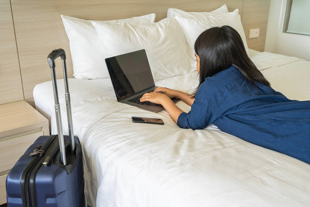 Asian woman lying on bed typing laptop next to suitcase - Фото, зображення