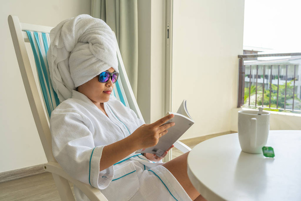 Young woman in bathrobe reading book in resort room - Фото, изображение