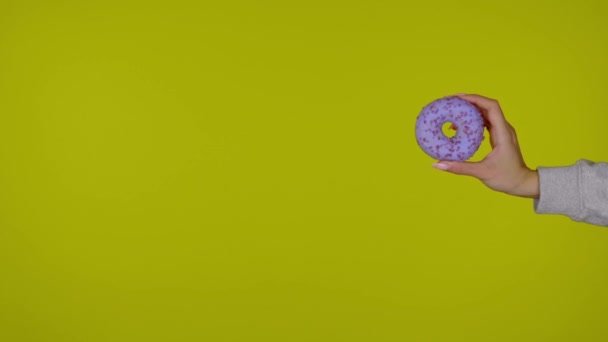 Vrouwelijke hand houdt een blauwe smakelijke en frisse donut vast, geïsoleerd over gele achtergrond met kopieerruimte. Reclamegebied, maak een model. Een close-up. 4k slow motion beelden - Video