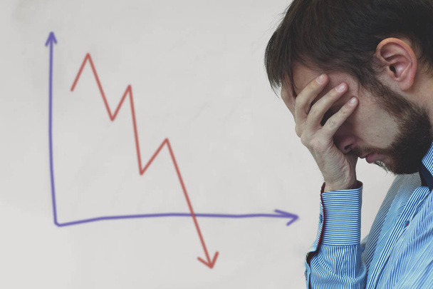 Frustrated business man standing near white board in office, covers his face with his hand at the falling graph of a stock market struck - 写真・画像