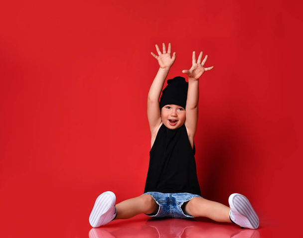 Small positive boy in stylish casual clothing, cap and white sneakers sitting on floor and feeling excited with hands raised up - Foto, immagini