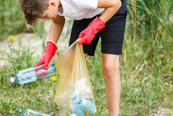 Boy in white t shirt in gloves collects garbage and plastic bottles into blue package on the beach. Young volunteer. Environmental protection, save environment concept. Close up hands in gloves - 写真・画像