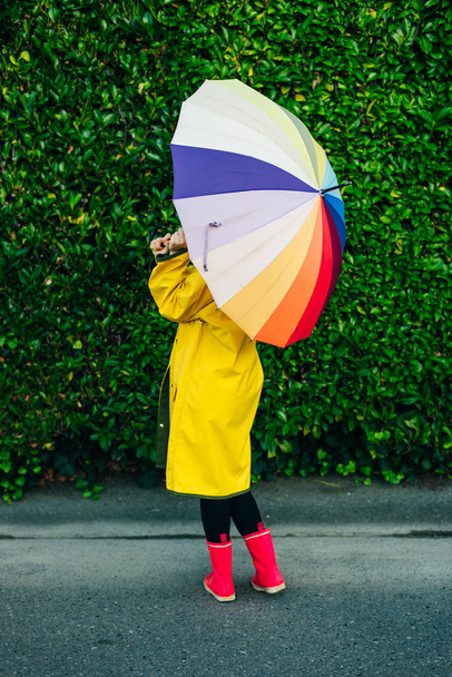 girl in a yellow raincoat on a background of a wall with grass with a multi-colored umbrella - Photo, Image