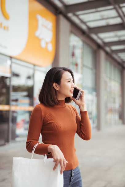 Muotokuva onnellinen nuori aasialainen nainen puhuu mobiili ja kuljettaa paperi ostoskassit kävely ulkona ostoskeskus
 - Valokuva, kuva