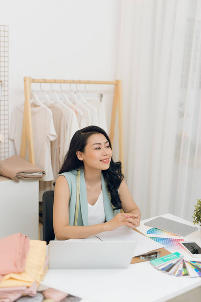 Portret van de jonge Aziatische mode-ontwerper zit aan het bureau voor haar laptop in de werkplaats - Foto, afbeelding
