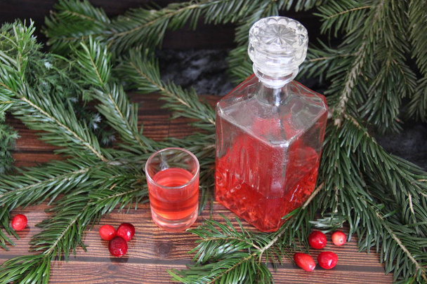 A bottle and a glass of cranberry tincture against a background of pine branches and berries on a dark table. Natural home medicine - Фото, изображение