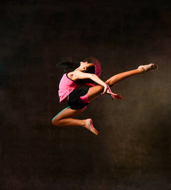 Young slim athletic girl gymnast dancer jumping up flying stretching doing gymnastic exercises in studio on dark wall - Foto, Imagem