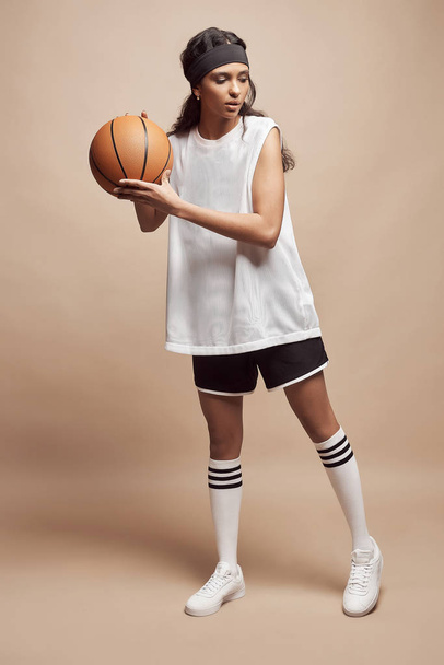 beautiful dark-skinned girl stands in full height on a beige background in white sports shirt, black shorts, white high socks and sneakers with a basketball ball and looks down - Photo, image