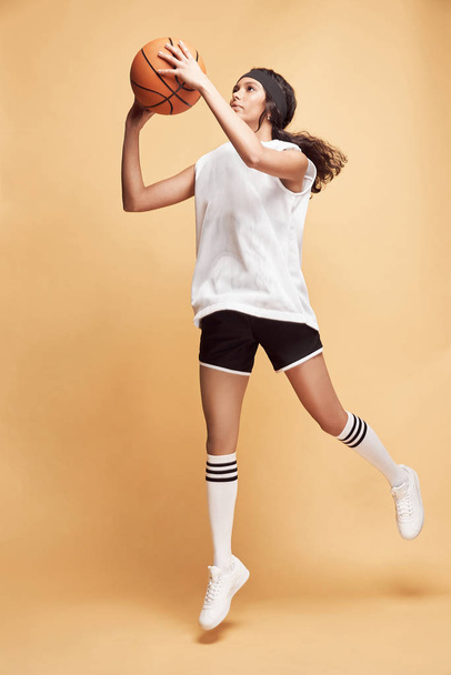 όμορφο σκουρόχρωμο κορίτσι σε κίτρινο φόντο με λευκό αθλητικό πουκάμισο, μαύρο σορτς, λευκές ψηλές κάλτσες και sneakers που χοροπηδούν με μπάλα μπάσκετ  - Φωτογραφία, εικόνα