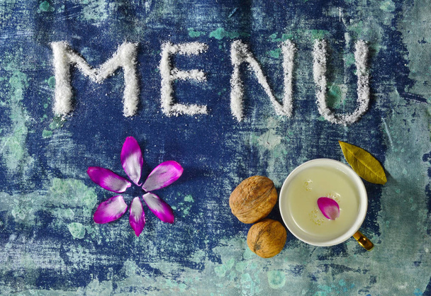 menu text made of salt with cup of milk, walnuts, bay leaves and petals isolated on grungy blue rugged background - Photo, Image