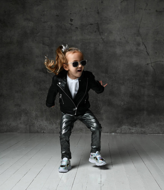 Happy excited kid girl in leather jacket, jeans and sunglasses is having fun jumping dancing screaming - Foto, imagen