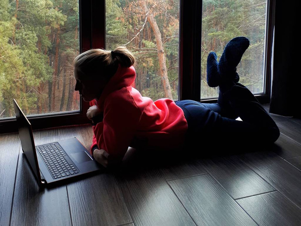молода чесна дівчина вдома працює на ноутбуці і розмовляє по телефону біля великого вікна
 - Фото, зображення